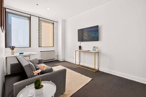 A modern living room with a grey sofa, a small white table with a plant, a large window, and a painting on the wall. - Stylish 1BR in the Heart of Auckland Zodiak Stays (Auckland)