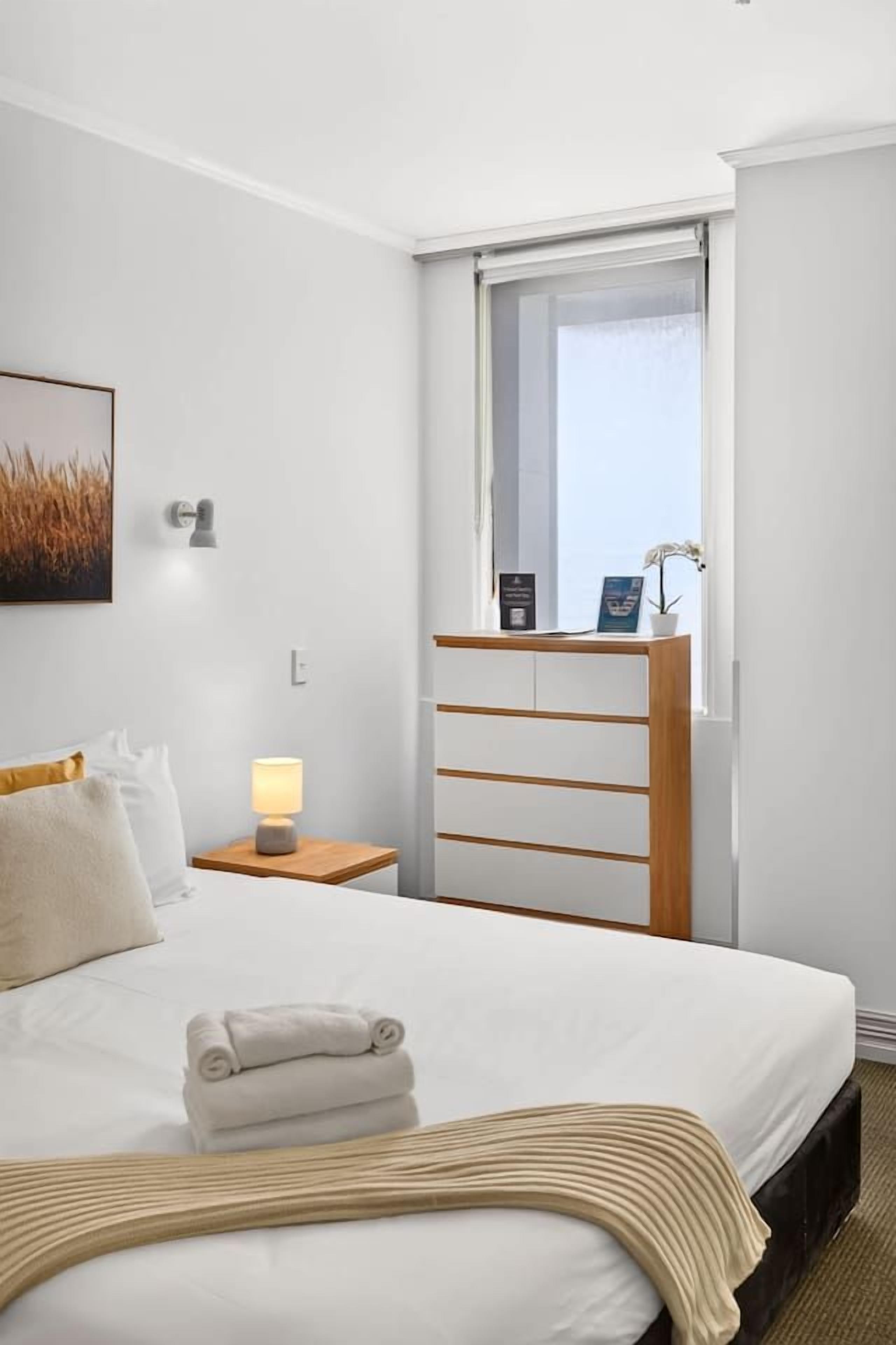 Et hotelværelse med en seng, et natbord, en lampe, et vindue og en vægmonteret hylde med en plante.