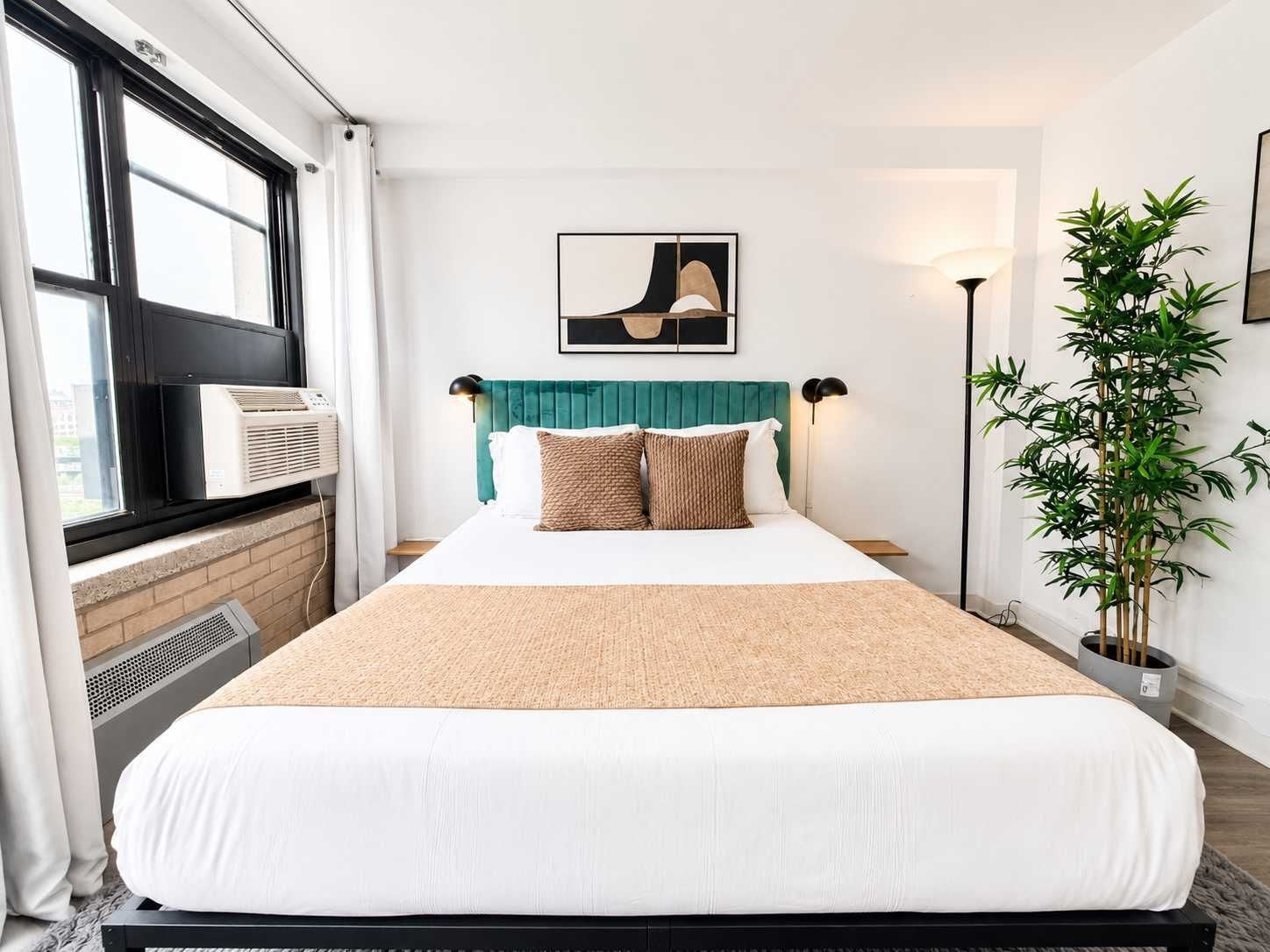 A hotel room with a bed, a window with curtains, a wall-mounted air conditioner, a floor lamp, a potted plant, and a framed picture on the wall.