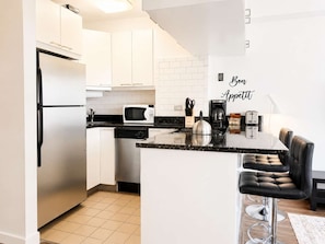 A modern kitchen with white cabinets, a microwave, a kettle, and a coffee maker. There is a bar stool and a sign that reads 'Bon Appetit.'