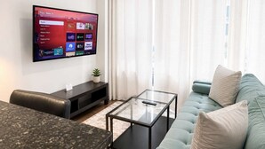 Una sala de estar con una televisión de pantalla plana montada en la pared, una mesa de centro de vidrio, un sofá y una planta pequeña sobre una consola.