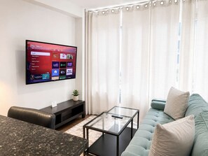 Uma sala de estar com televisão de tela plana na parede, mesa de centro de vidro, sofá e uma pequena planta em um aparador.