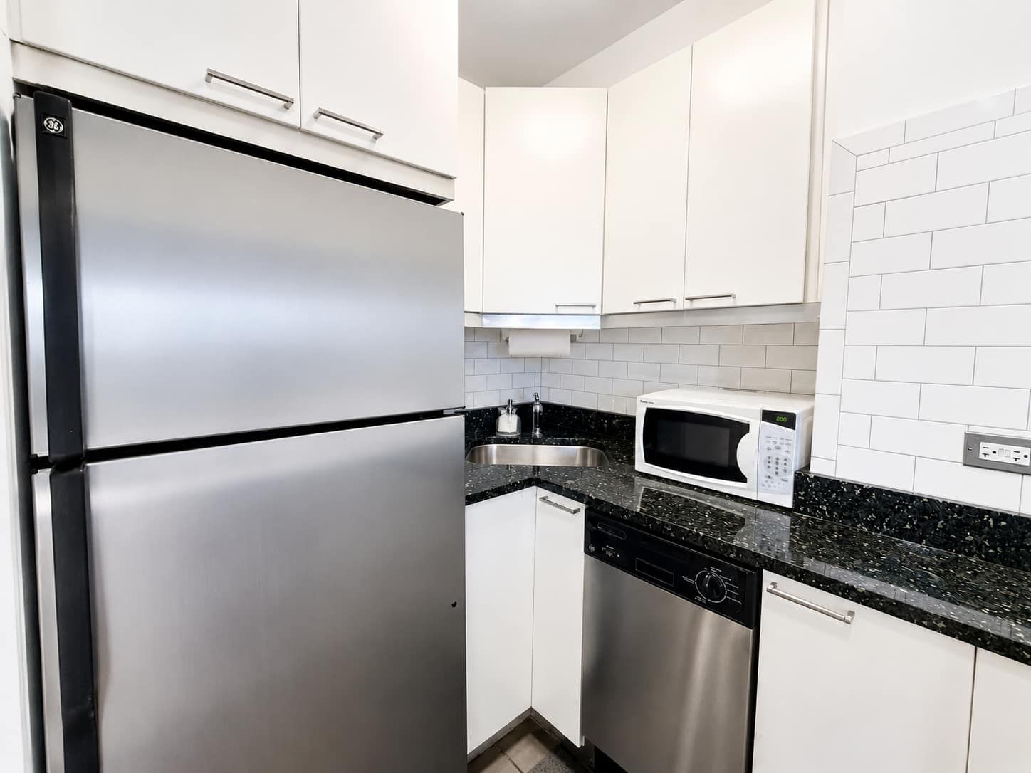 Cozinha moderna com eletrodomésticos de aço inoxidável, armários brancos e bancada de granito preto.