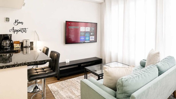 Un salón moderno con un televisor de pantalla plana montado en la pared, un sofá, una mesa de centro y una zona de comedor con un taburete de bar.