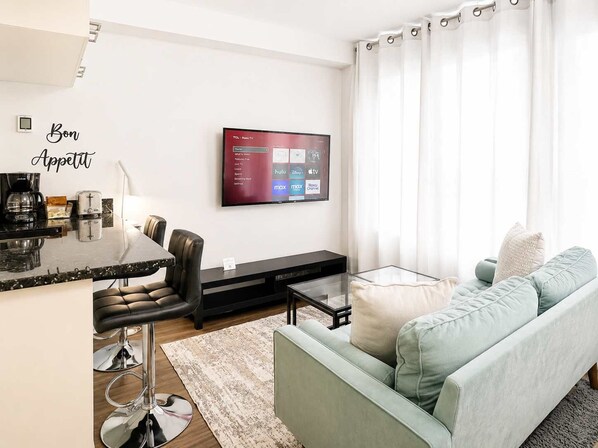 Uma sala de estar moderna com televisão de tela plana na parede, um sofá, uma mesa de centro e um espaço de jantar com um banco alto.
