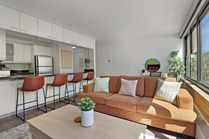 Un espacio habitable moderno con un conjunto de sofás, una mesa de centro con una planta y una zona de cocina con taburetes de bar.