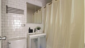 Une salle de bain avec des murs recouverts de carreaux blancs, des toilettes, un lavabo avec un miroir au-dessus et un rideau de douche.