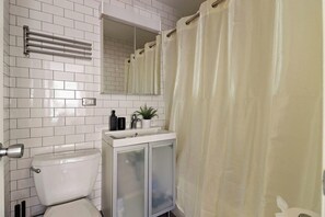 Un baño con paredes de azulejos blancos, un inodoro, un lavabo con un espejo encima y una cortina de ducha.
