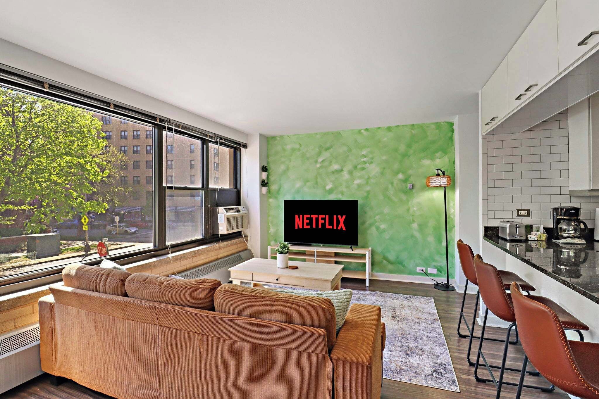 A modern living room with a large window, a green accent wall, a sofa, a coffee table, and a kitchen area with a sink and countertop.