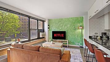 A modern living room with a large window, a green accent wall, a sofa, a coffee table, and a kitchen area with a sink and countertop.