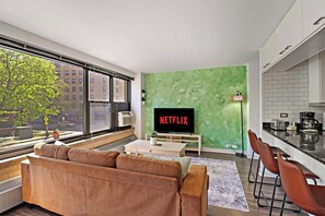 A modern living room with a large window, a green accent wall, a sofa, a coffee table, and a kitchen area with a sink and countertop.