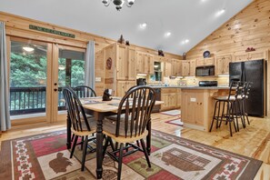 A cabin interior with a dining area, kitchen, and a large window with a view of trees. - Family Getaway Hot Tub, Fire Pit, Game Room, Pool, Gym, near apple orchards (Ellijay)