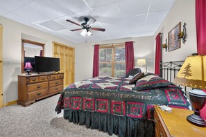 Une chambre à coucher comprenant un lit, une commode, un téléviseur, un ventilateur de plafond, une fenêtre avec des rideaux et une lampe.