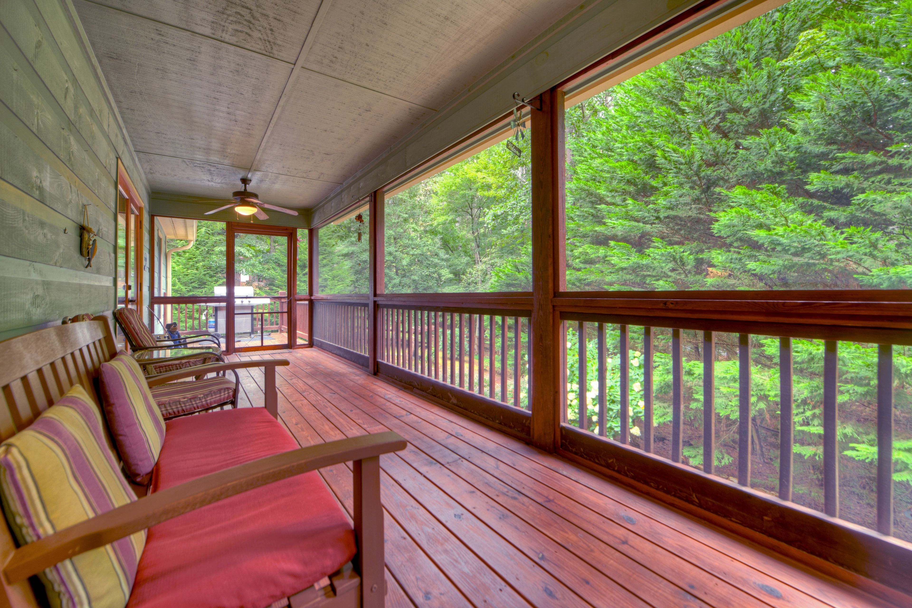 A wooden porch with a bench and a ceiling fan, overlooking a forested area.