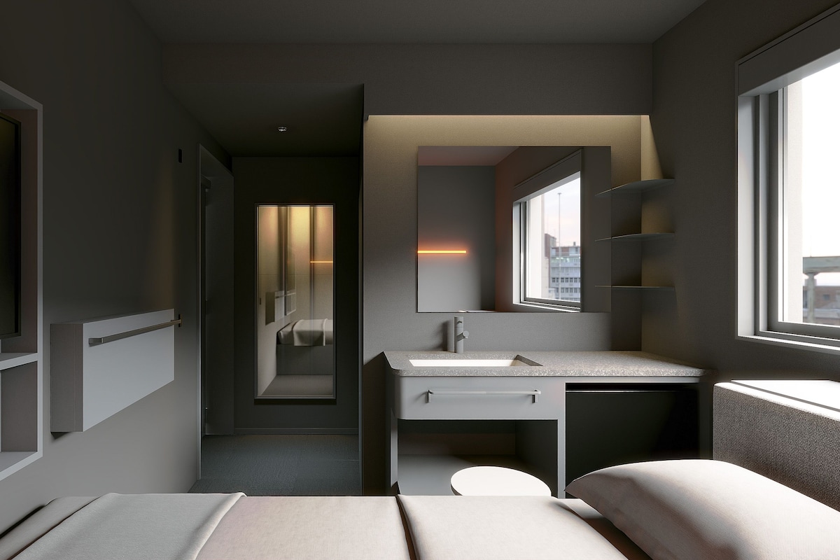 A modern hotel room with a bed, a bedside table, a mirror, a window, and a bathroom with a sink and toilet.