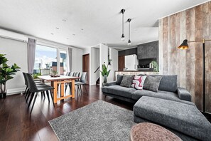 En moderne stue med spiseområde, en grå sofa, et sofabord og et køkken, der kan ses gennem en åben dør.
