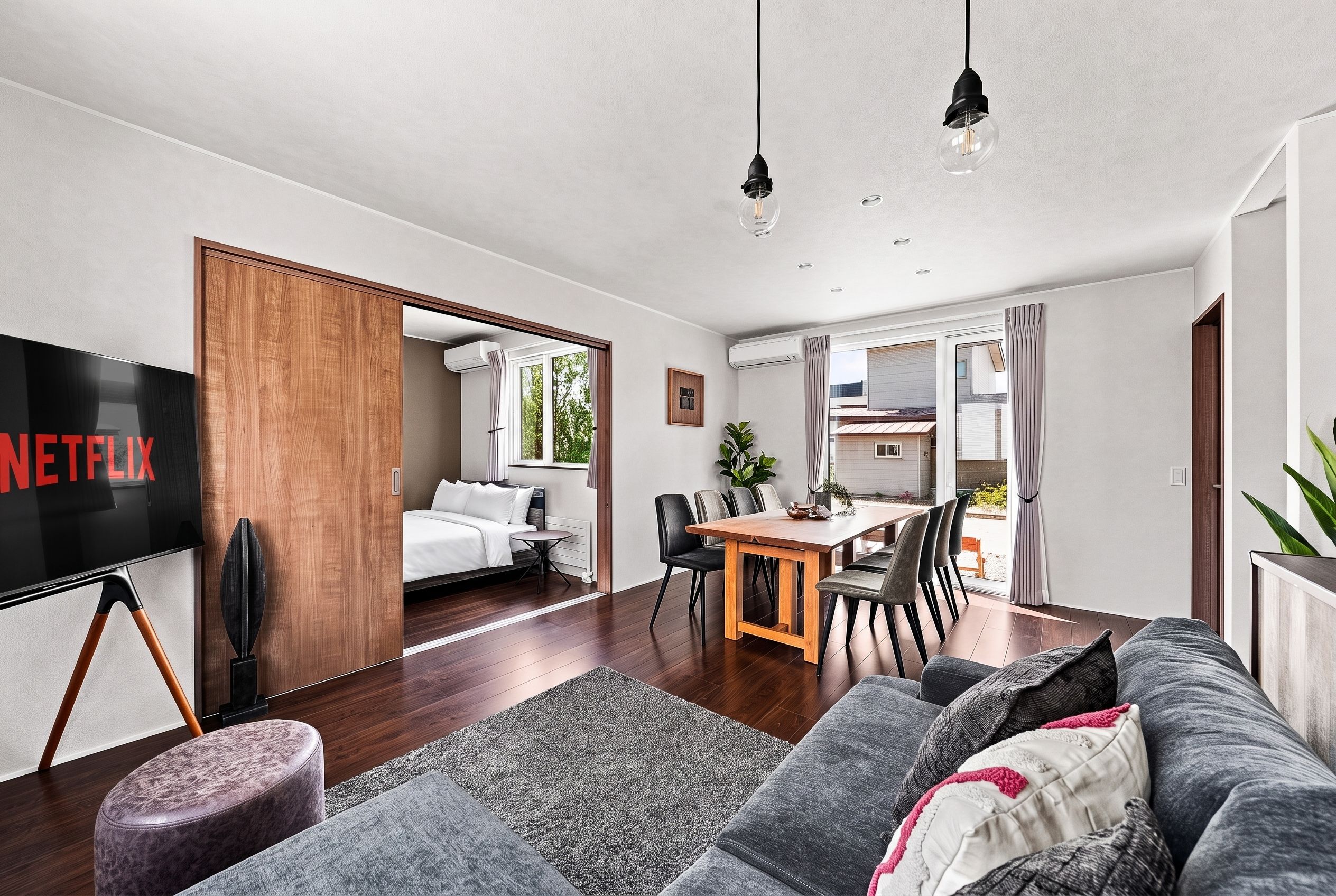 A modern living room with a flat-screen TV, a sofa, a dining area with chairs, and a kitchen visible through an open door.