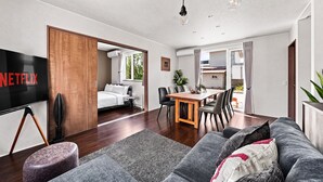 A modern living room with a flat-screen TV, a sofa, a dining area with chairs, and a kitchen visible through an open door. - The Cross (Asahikawa)