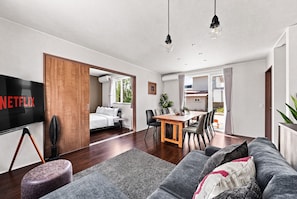Uma sala de estar moderna com televisão de tela plana, um sofá, uma área de jantar com cadeiras e uma cozinha visível por uma porta aberta.