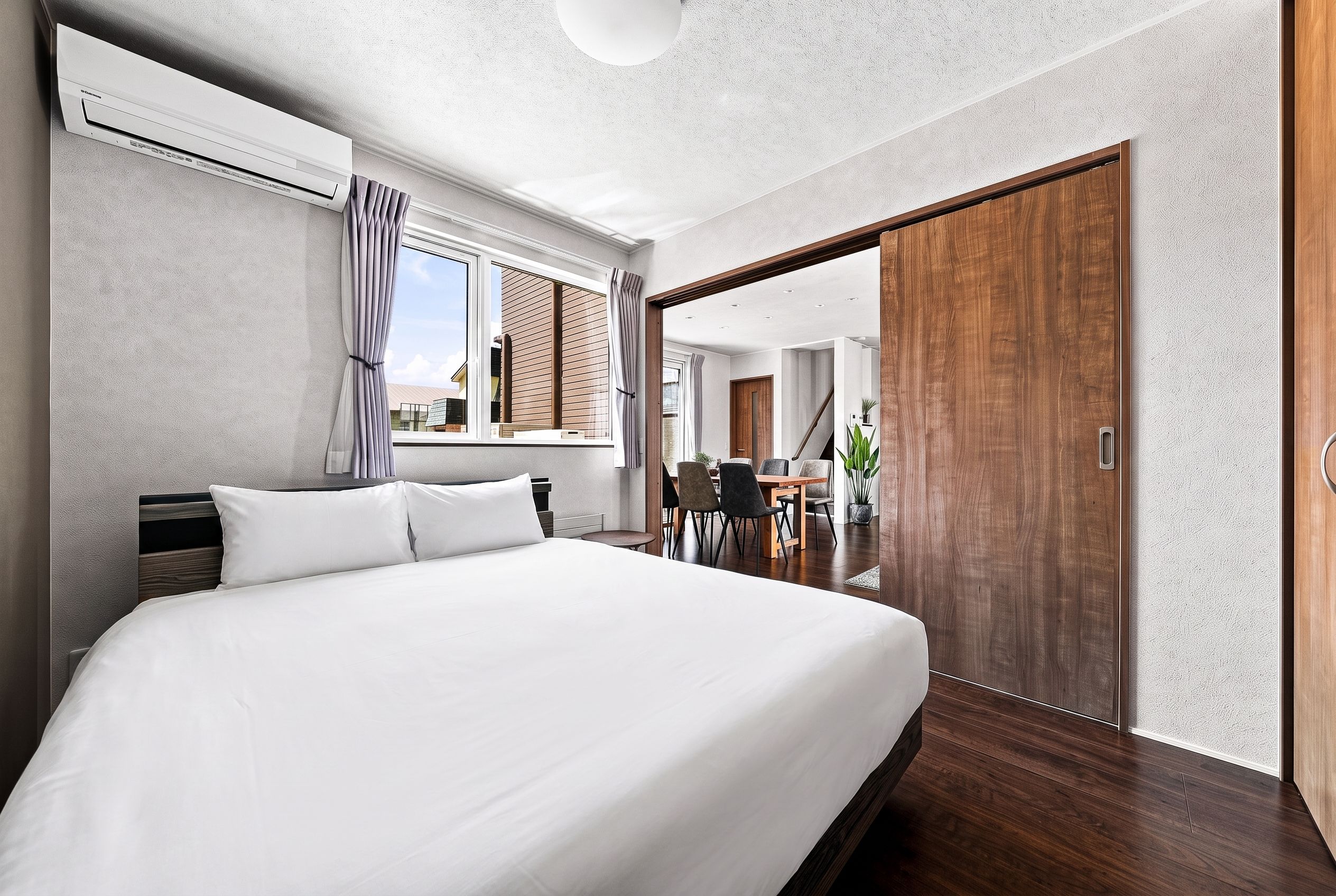 A modern hotel room with a large bed, a window with curtains, a wooden door, and a view of the outside.