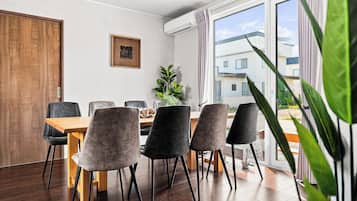 Un espace de repas moderne avec une table et des chaises en bois, une plante en pot et une vue sur les bâtiments à travers de grandes fenêtres.
