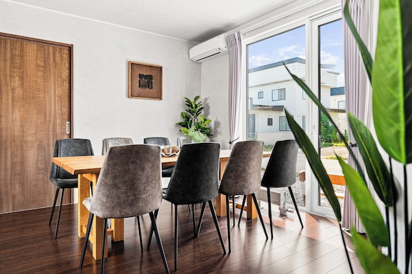 Un comedor moderno con una mesa y sillas de madera, una planta en maceta y vistas a edificios a través de amplios ventanales.