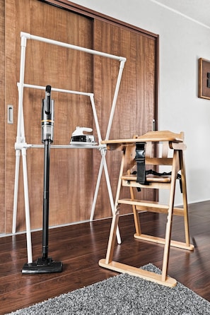 Habitación con piso de madera, un ventilador de pie, una silla de madera y una estructura de tuberías blanca contra una pared de madera.