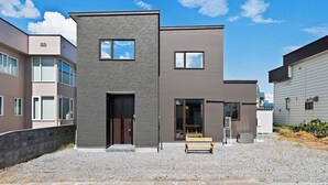 A modern two-story building with a flat roof, large windows, and a gravel driveway. - The Cross (Asahikawa)