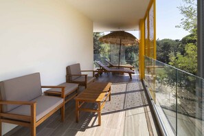 A balcony with wooden furniture, a thatched umbrella, and a glass railing overlooking a forested area. - Holiday flat with air conditioning (Bibione)