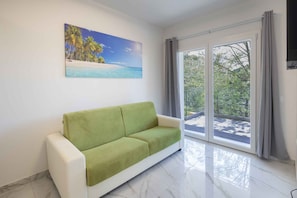 Ein Zimmer mit einem grünen Sofa, einem Strandbild und einem großen Fenster mit Vorhängen.