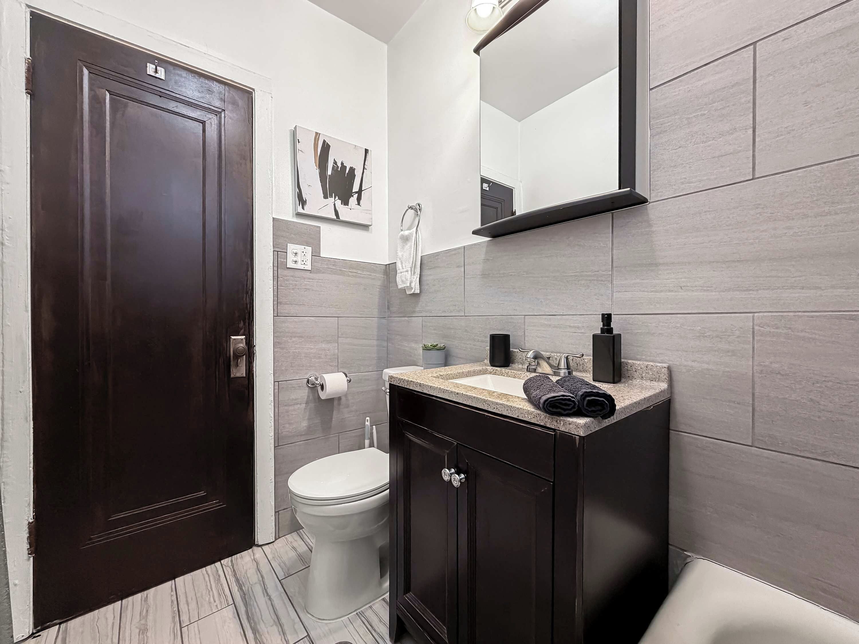 Ein Badezimmer mit einer dunkelholznen Tür, einer weißen Toilette, einem Waschbecken mit einer Granit-Arbeitsplatte und einem Spiegel über dem Waschbecken.