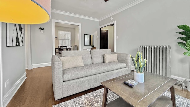 Un salon avec un canapé gris, une table basse en bois, un ventilateur de plafond et un radiateur.