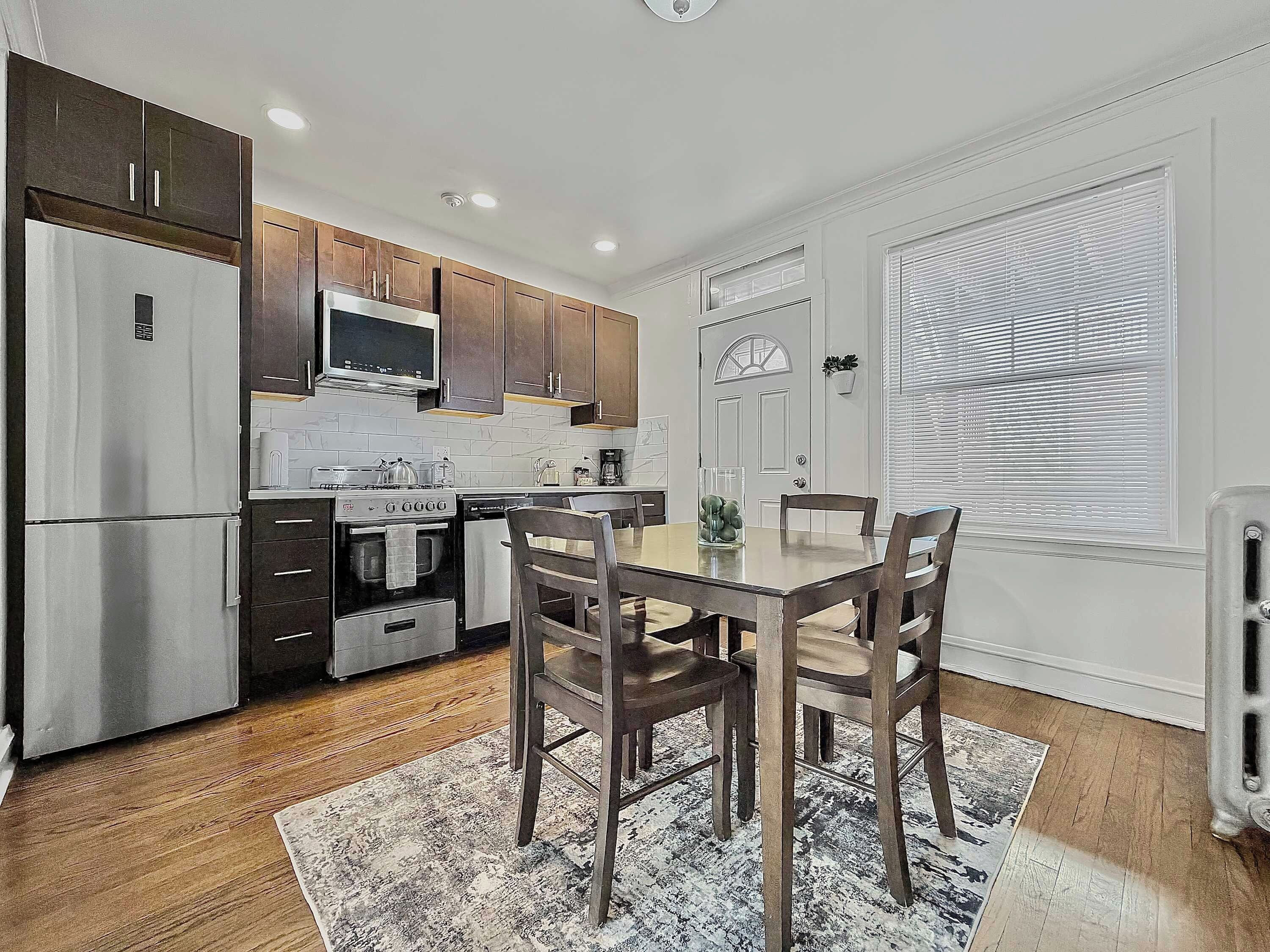A modern kitchen with stainless steel appliances, wooden cabinets, a dining table with chairs, and a large window with blinds.