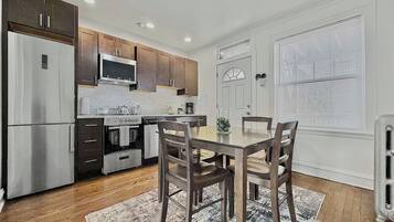 A modern kitchen with stainless steel appliances, wooden cabinets, a dining table with chairs, and a large window with blinds.