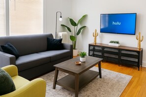 A living room with a sofa, a coffee table, a television displaying the Hulu logo, a plant, and a cactus on a side table.