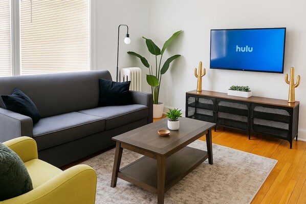 A living room with a sofa, a coffee table, a television displaying the Hulu logo, a plant, and a cactus on a side table. - 1BR Unit w/ 2 Comfy Beds (Chicago)