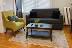 A living room with a sofa, a chair, a coffee table, a plant, and a radiator. - 1BR Unit w/ 2 Comfy Beds (Chicago)