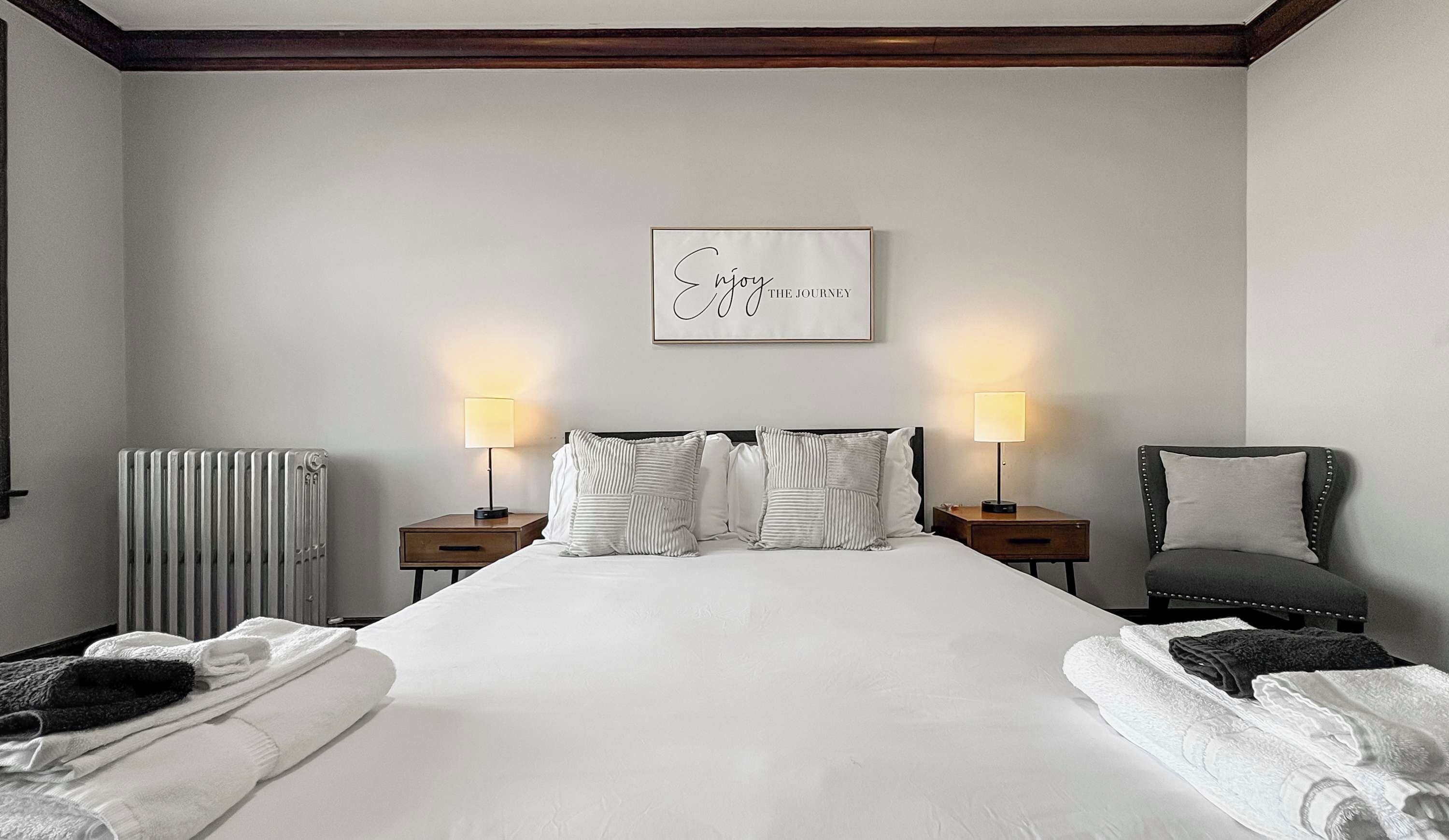 Un lit bien fait, avec du linge de lit blanc, deux tables de chevet avec des lampes, un radiateur et une citation encadrée accrochée au mur.