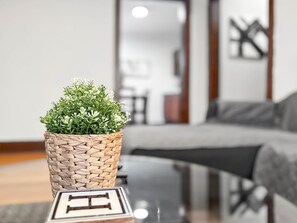 A potted plant on a table with a letter 'H' in front of it, in a room with a gray sofa and a mirror on the wall.