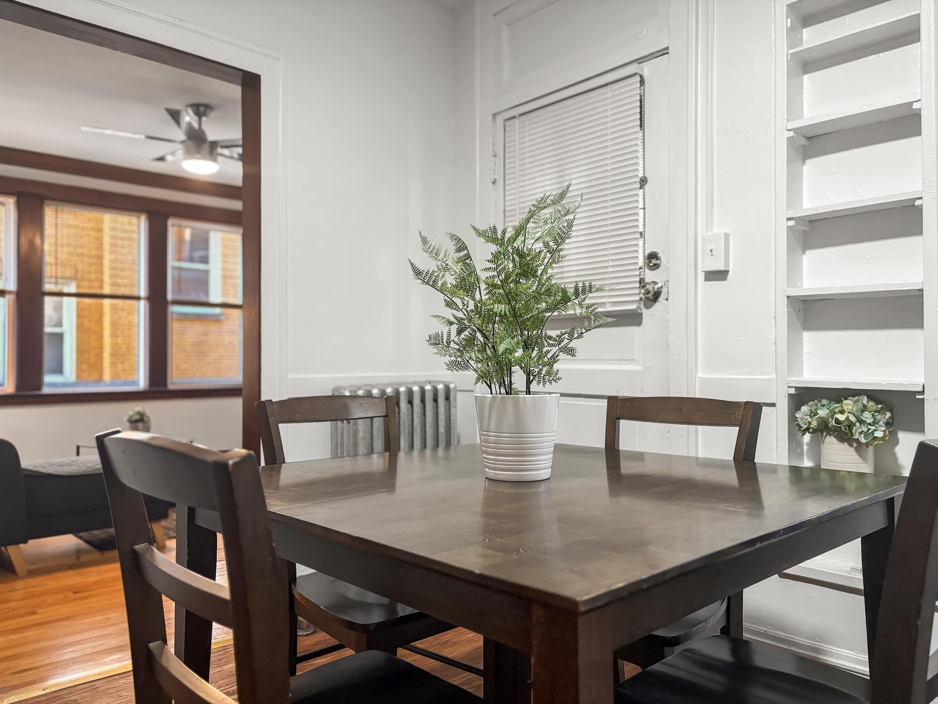 Een eetruimte met een houten tafel, vier stoelen, een potplant met een varen en een radiator.