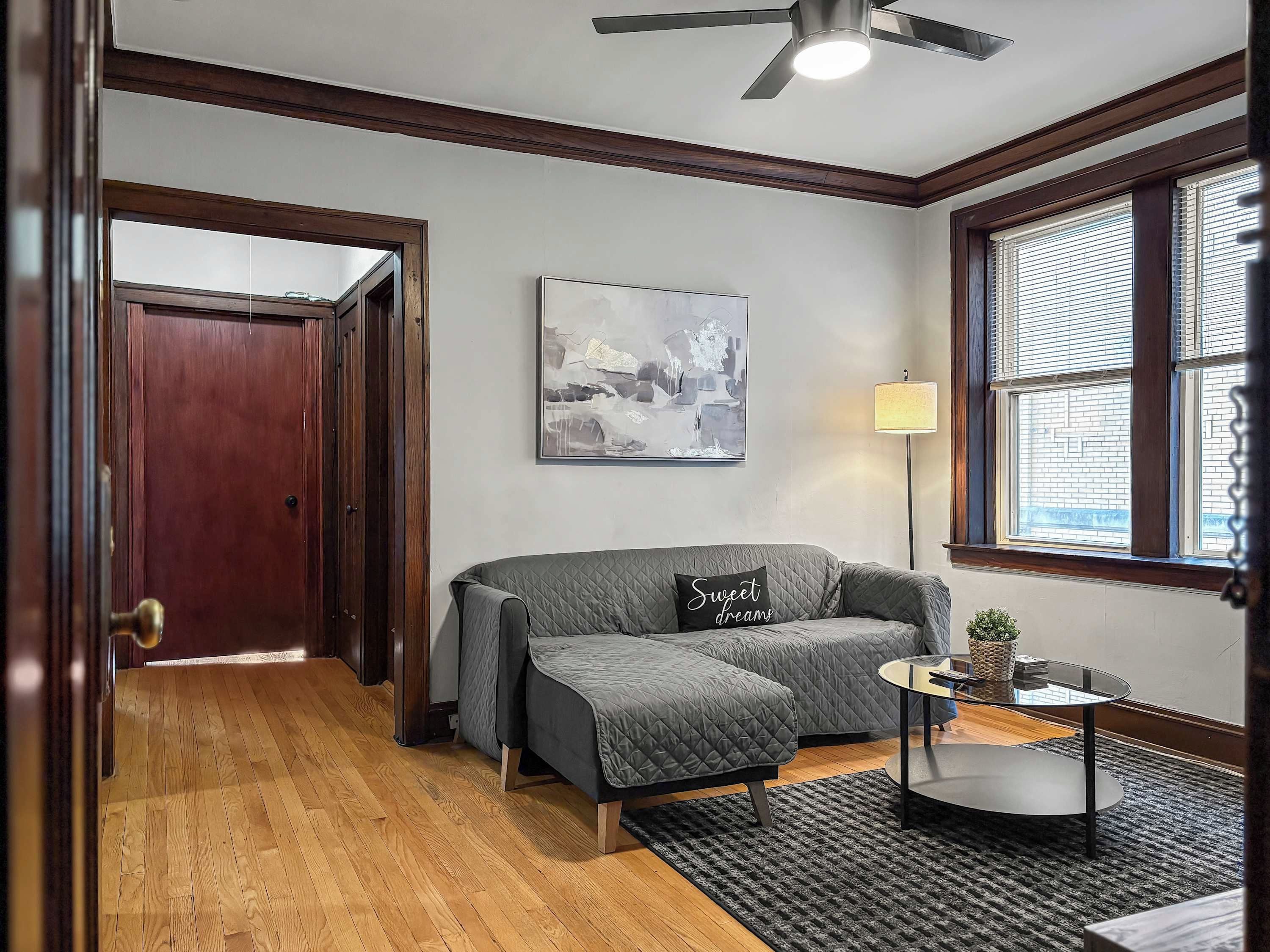 A room with a grey sofa, a painting on the wall, a lamp, and a window with blinds.