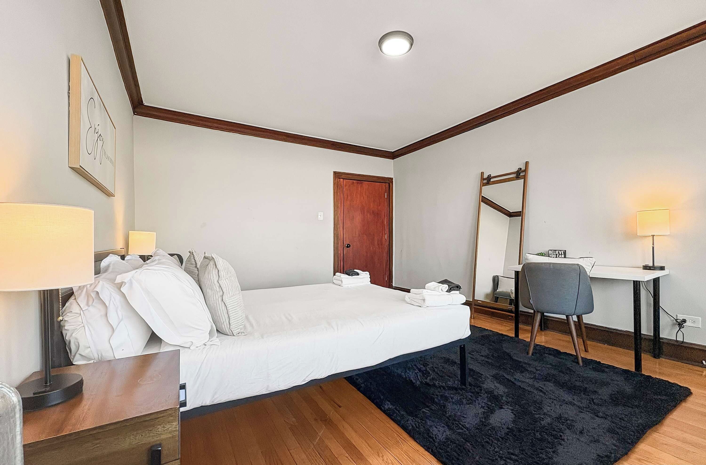 A hotel room with a bed, bedside table, desk, chair, and a black rug.
