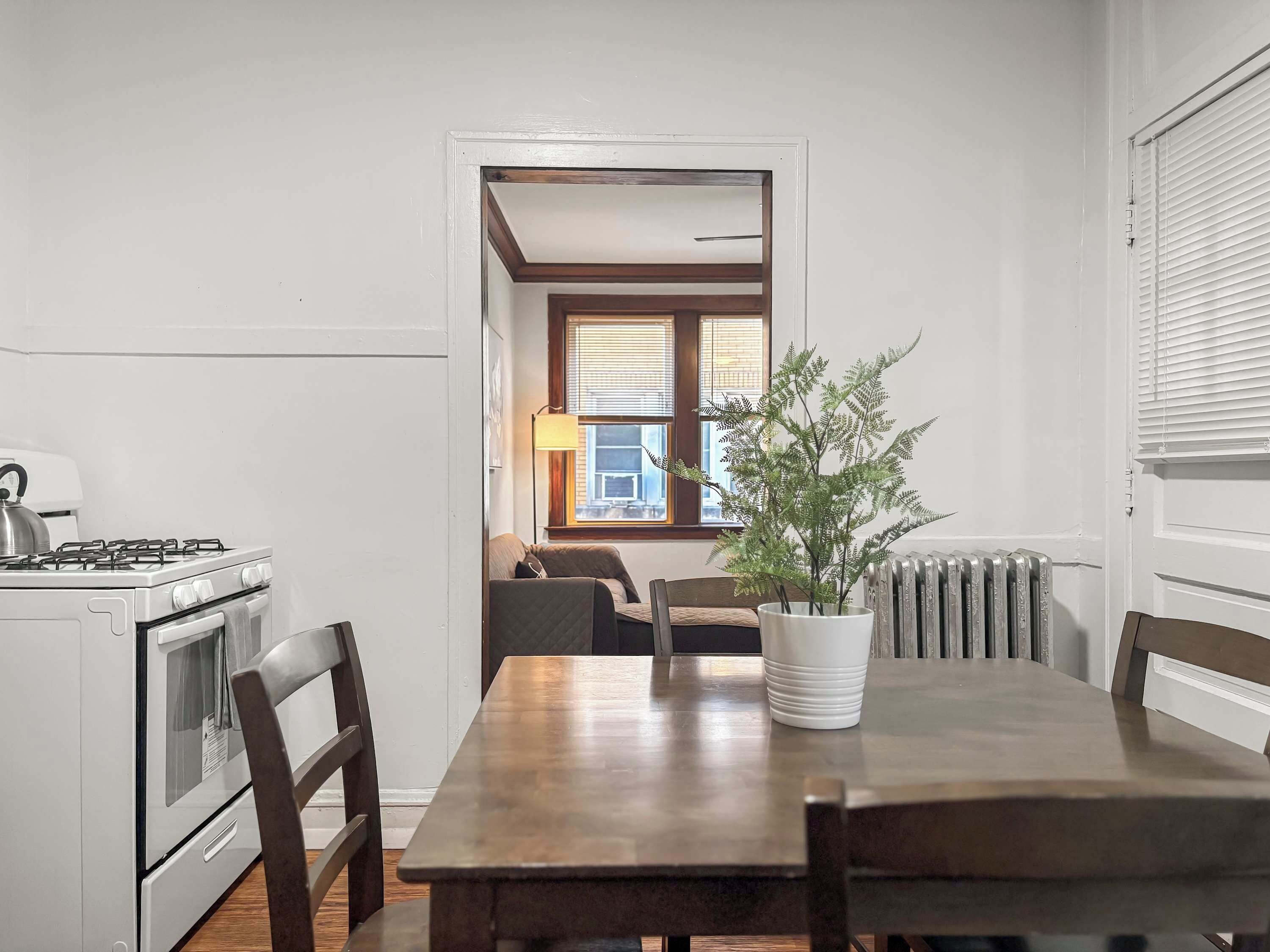 Een keuken met een fornuis, oven en een eettafel met stoelen. Op tafel staat een potplant en er is een raam met jaloezieën op de achtergrond.