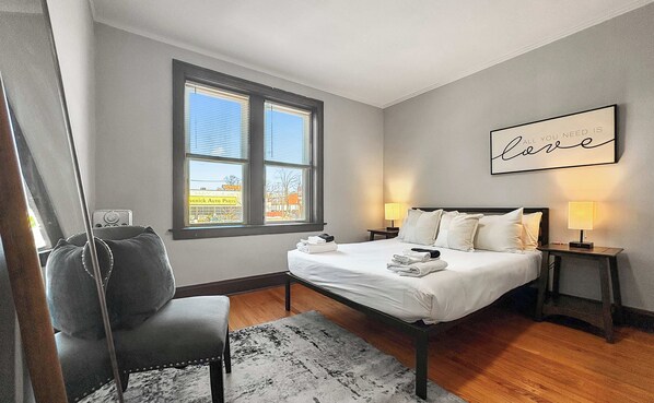 A bedroom with a bed, two bedside lamps, a chair, a rug, a window with blinds, and a wall with a framed quote.