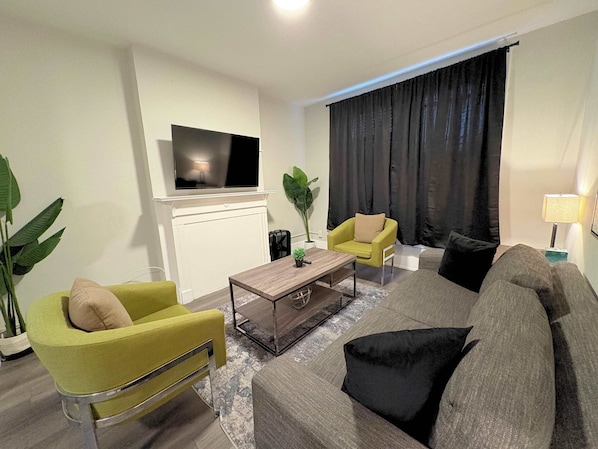 A modern living room with a flat-screen TV mounted on the wall, a coffee table, two armchairs, a sofa, and a large window with dark curtains. - Comfortably Appointed & Spacious 3BR Apartment (Chicago)