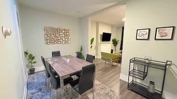 Uma área de refeições com uma mesa de madeira, cadeiras pretas e um tapete cinzento. Há um espelho fixado na parede e duas obras de arte emolduradas.