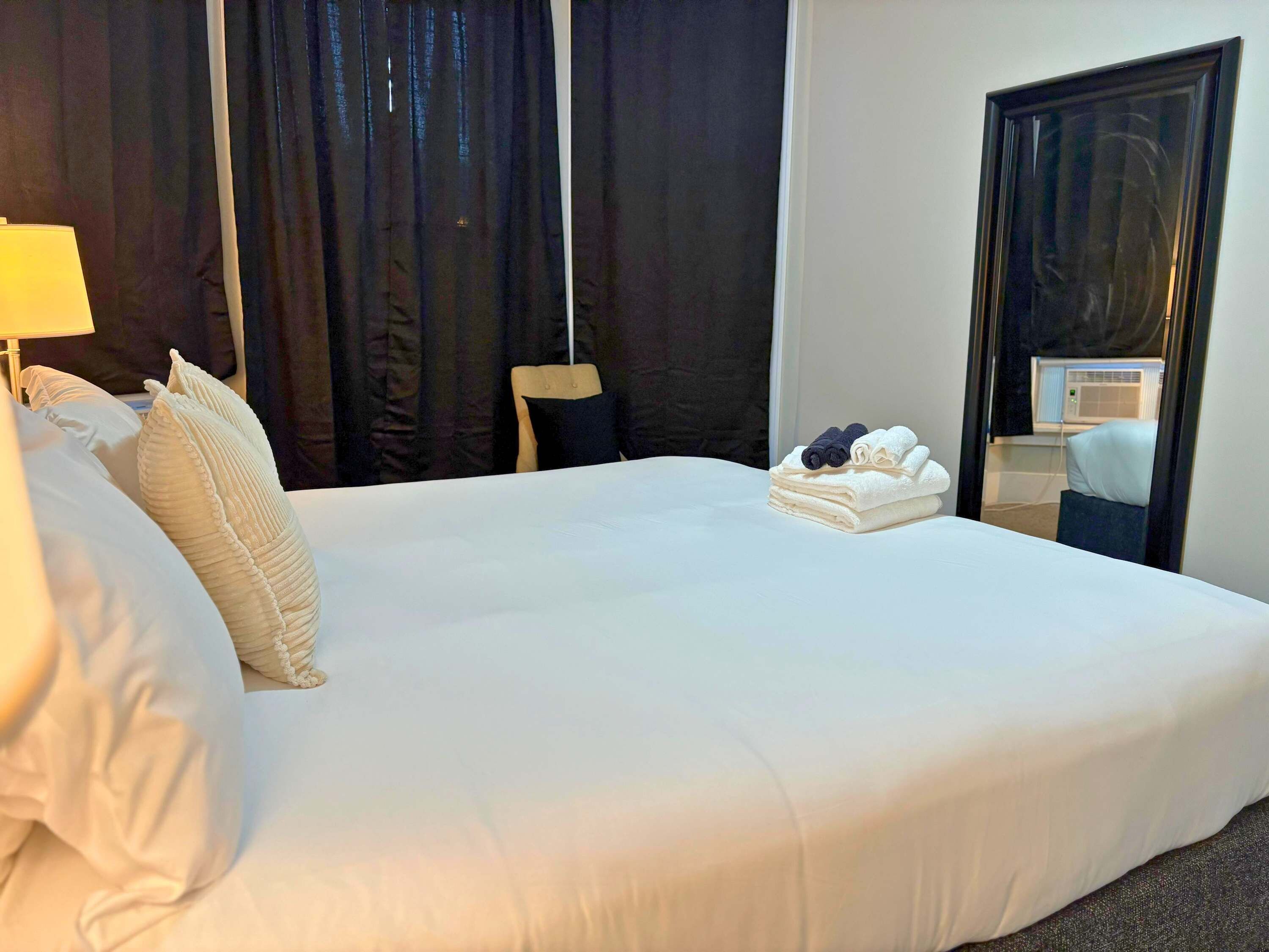 Cama bem arrumada com lençóis brancos, uma pilha de toalhas dobradas e um espelho refletindo parte do quarto.
