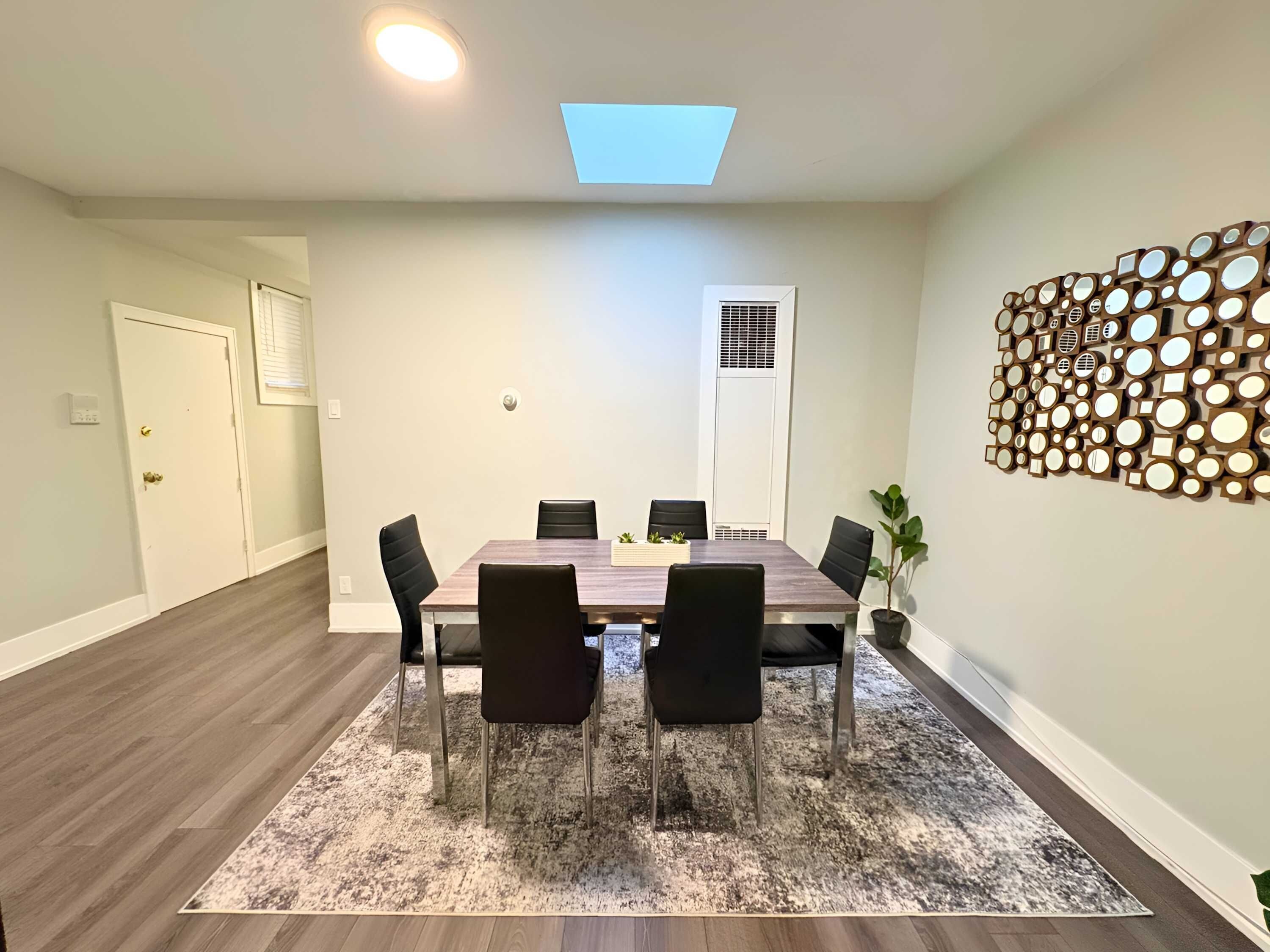 Uma área de jantar com uma mesa retangular, seis cadeiras, um tapete estampado, uma luminária no teto, um respiradouro e um objeto decorativo fixado na parede.