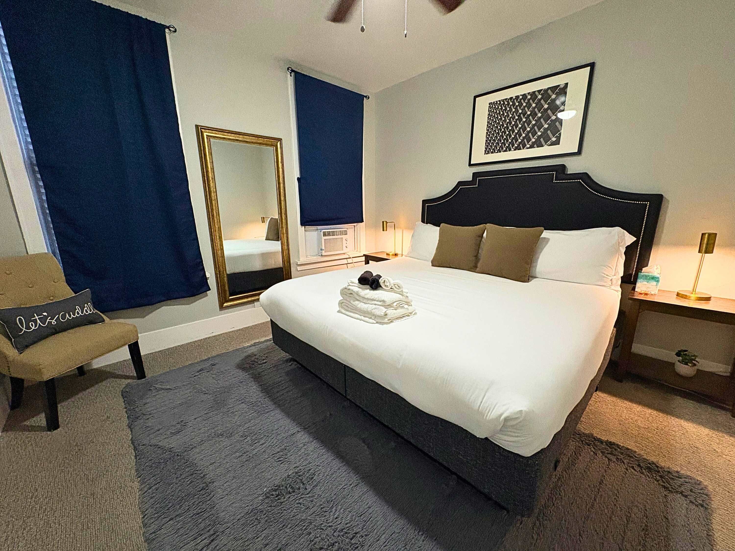 A hotel room with a large bed, a chair, a mirror, a bedside table with a lamp, and a framed picture on the wall.