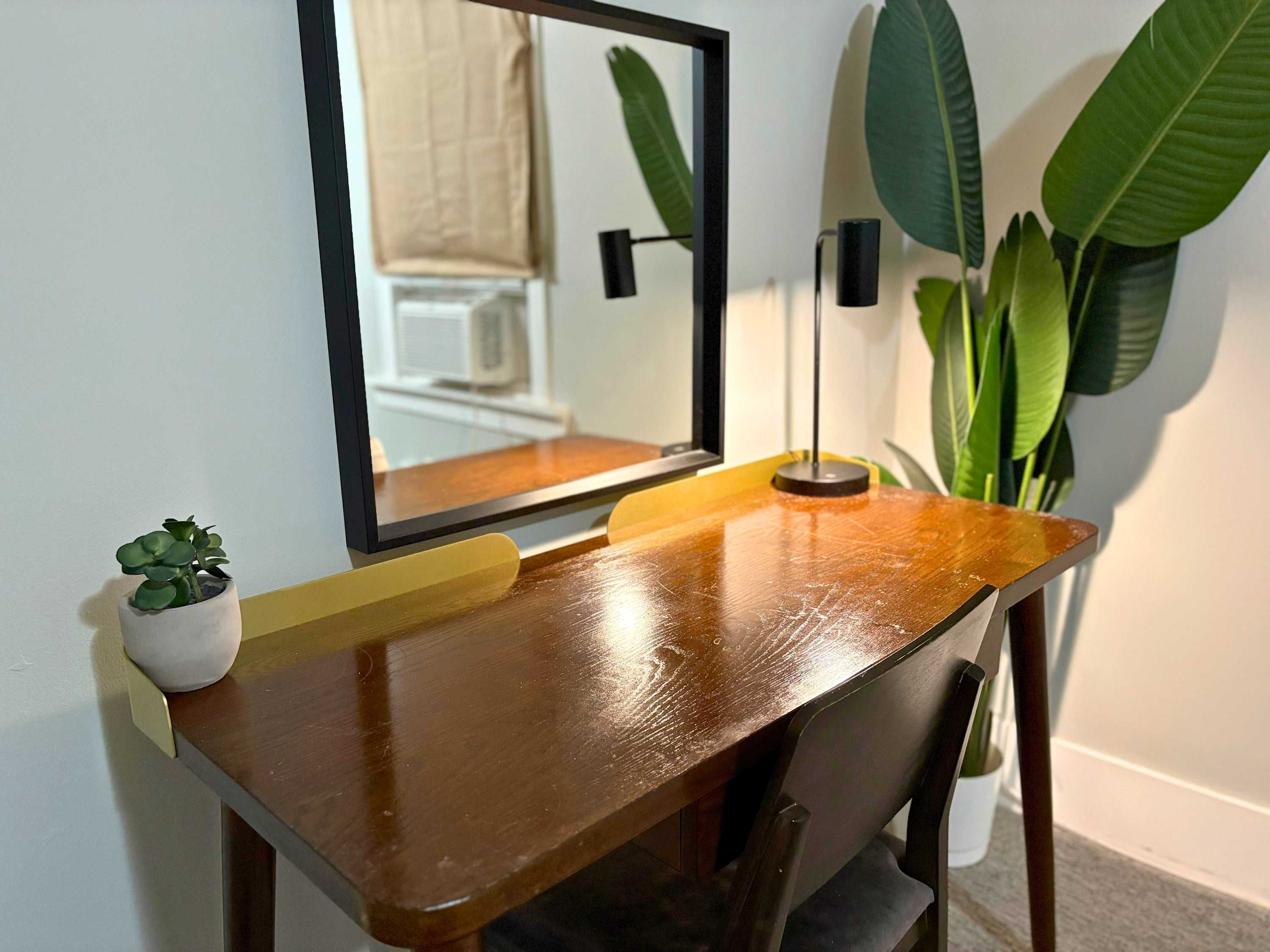 Uma mesa de madeira com um espelho, uma pequena planta em vaso e uma luminária.